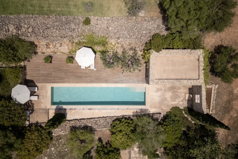 Toma de dron de una piscina minimalista con escaleras de entrada, rodeada por una cubierta de madera y gruesos muros de piedra rústica. Hay vegetación en los bordes y dos sombrillas blancas.