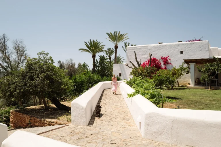 Un sendero de piedra flanqueado por muros blancos conduce a una casa de estilo mediterráneo. Una mujer con vestido rosa camina junto a un perro oscuro. Alrededor hay palmeras, arbustos verdes y buganvillas rojas.