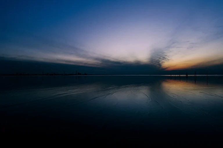 Cielo nocturno azul oscuro que se fusiona con el horizonte en un brillo naranja. El agua oscura refleja los colores del cielo. Se distingue la tenue silueta de una línea de edificios o fábricas.