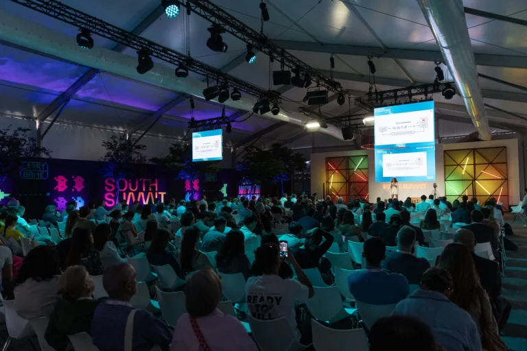 Toma amplia de un gran auditorio lleno de gente en un evento tecnológico (South Summit). Una ponente está en el escenario bajo focos y grandes pantallas laterales que muestran la presentación.