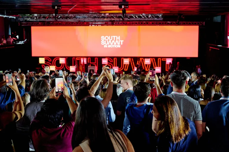Vista trasera de una gran audiencia en una conferencia o cumbre, donde muchas personas graban el escenario y la pantalla principal con el logo de "South Summit" en color naranja brillante.
