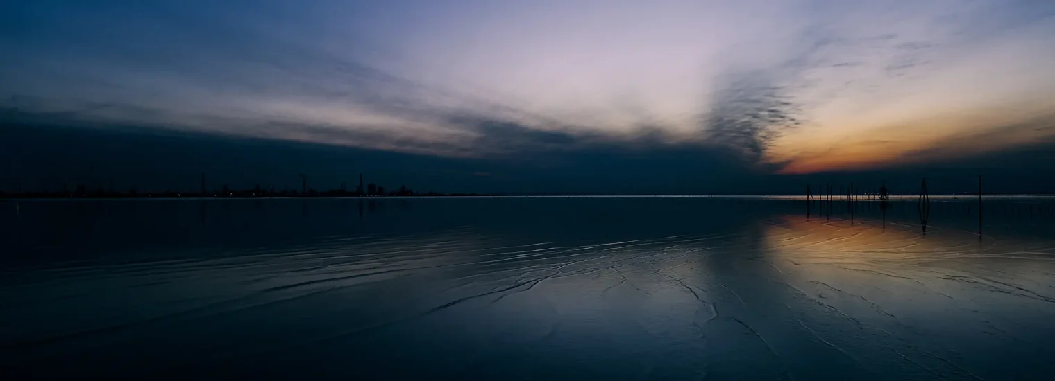 Cielo nocturno azul oscuro que se fusiona con el horizonte en un brillo naranja. El agua oscura refleja los colores del cielo. Se distingue la tenue silueta de una línea de edificios o fábricas.