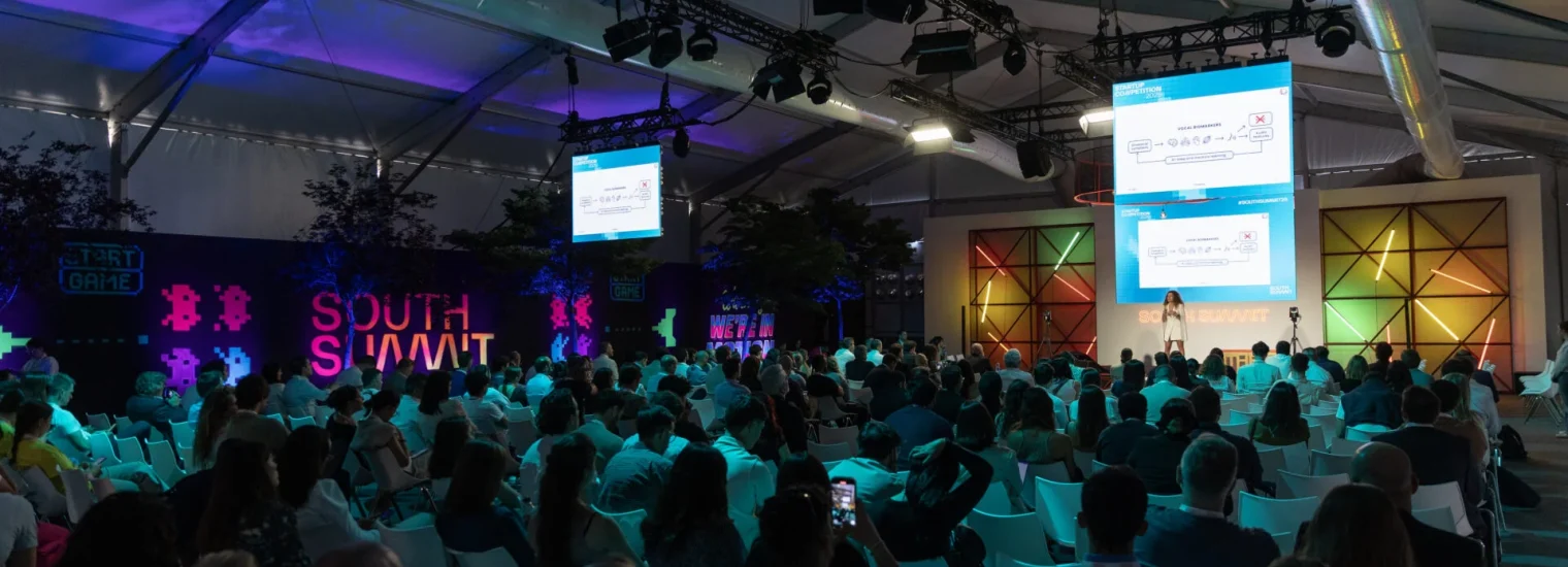 Toma amplia de un gran auditorio lleno de gente en un evento tecnológico (South Summit). Una ponente está en el escenario bajo focos y grandes pantallas laterales que muestran la presentación.